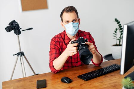 Freelancer photographer, videographer works using a medical mask from home during a pandemic, epidemic of coronavirus, seasonal flu. Young man uses computer for processing, photo camera in his handsの写真素材
