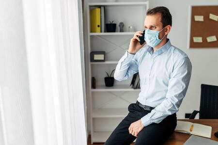 Stay at home. A young man works from home, home office, work remotely. Medical masked businessman talking on cell phoneの写真素材