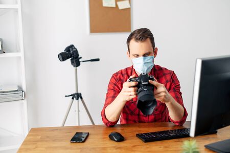 Freelancer photographer, videographer works using a medical mask from home during a pandemic, epidemic of coronavirus, seasonal flu. Young man uses computer for processing, photo camera in his handsの写真素材