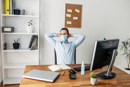 The man stayed at home and works remotely. The guy with the medical mask takes a break and leaned back in his chair relaxed. Coronavirus Pandemic, Flu, Covid 19の写真素材