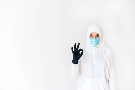 A woman in a white protective suit and medical mask stands with a hand in a rubber glove, showing the sign OK, control the situation. Isolated on a white background. The concept of resisting the virusの写真素材