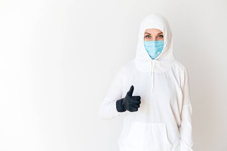 A woman in a white protective suit and medical mask stands with a hand in a rubber glove, showing the sign thumb up, control the situation. White background. The concept of resisting the virusの写真素材