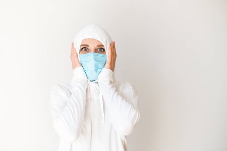 Quarantine, epidemic, pandemia. A woman in a white protective suit and a medical mask with her scared eyes wide open. She holds her head in her hands. Isolated on a white backgroundの写真素材