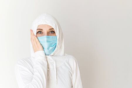 Quarantine, epidemic, pandemia. A woman in a white protective suit and a medical mask with her scared eyes wide open. She holds her hand near face. Isolated on a white backgroundの写真素材
