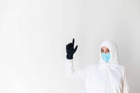 Woman in a white protective suit and medical mask with white background. She is standing with her hand up in a rubber glove showing a good idea sign. Coronavirus control conceptsの写真素材