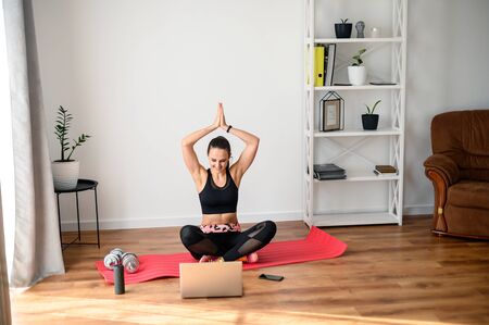 Online training, online video tutorial. A young woman in sportswear is using a laptop to watching yoga video tutorial at home. She sits on the mat, looks at the screen and repeats the exercises.の写真素材