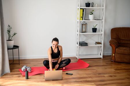 Get in shape at home. A young girl uses a workout video tutorial, she sits on a mat in the room, does exercises and looks at the laptop. Selfdissipline and healthy lifestyle conceptの写真素材