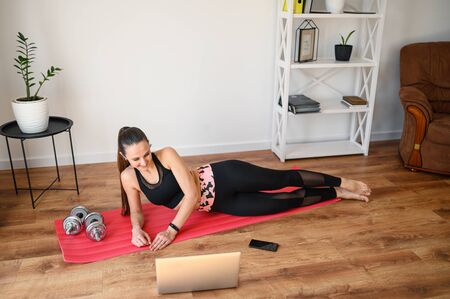 Online training, online video tutorial. A young woman in sportswear is using a laptop to watching workout video tutorial at home. She lying on the mat, looks at the screen and repeats the exercises.の写真素材