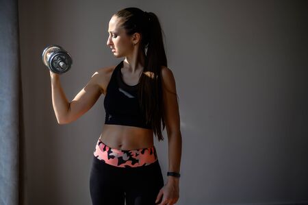 Doing sports at home during quarantine. Get in shape. Attractive athletic woman is doing workout at home. She in a trendy sportswear lifts a dumbbellの写真素材
