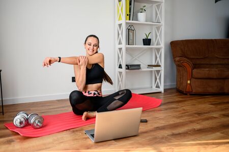 Get in shape at home. A young girl uses a workout video tutorial, she sits on a mat in the room, does exercises and looks at the laptop. Selfdissipline and healthy lifestyle conceptの写真素材