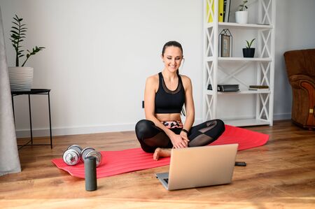 Online training, online video tutorial. A young woman in sportswear is using a laptop to watching workout video tutorial at home. She sits on the mat and looks at the screenの写真素材