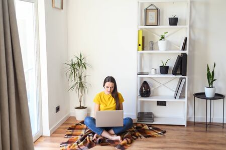 Freelancer woman works from home, work remotely. Young woman in casual clothes sitting with a laptop on a cozy plaid in a stylish minimalistic interior.の写真素材