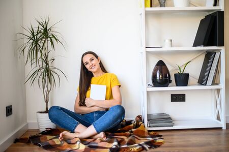 A woman spends time reading books. Positive attractive woman sitting on a cute plaid with a book in her hands, she is looking at the camera and smilingの写真素材