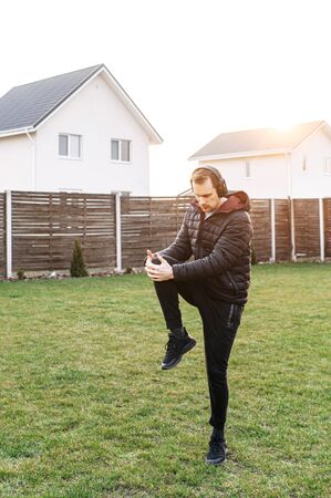 Self isolation. Workout in the backyard. A young guy does a warm-up, exercises in the backyard on the lawn. He is in the headphonesの写真素材