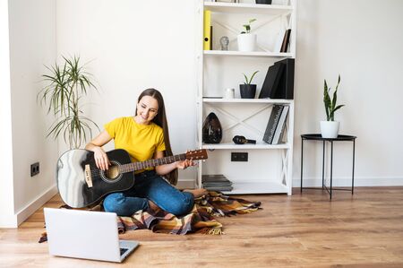 Online training, online classes. A young woman watches video tutorial on guitar playing, she sits on a cozy plaid with a guitarの写真素材