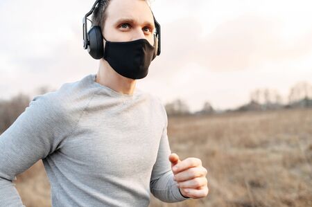 Sport during quarantine, self-isolation in the countryside. A young athletic guy is jogging on a dirt road in the meadow. He is wearing a black medical mask and headphonesの写真素材