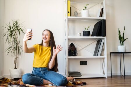 Phone video call. A young woman communicates via video, zoom. She is sitting on the floor in a Scandinavian style room.の写真素材