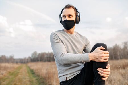 Protect yourself during a pandemic. Sports in a medical mask. Young atletic guy in headphones doing outdoor workout. Closeup portrait in sunset lightの写真素材