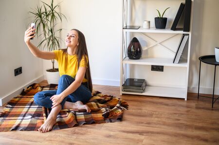 Selfie time. Young attractive girl in a yellow T-shirt takes a selfie on the phone in a cozy home atmosphereの写真素材