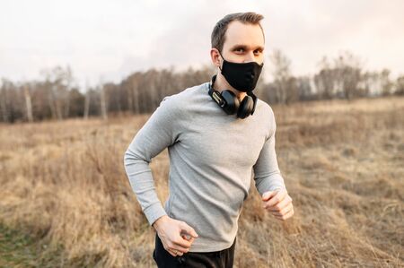 Sports during the quarantine period. A young guy in a black protective medical mask is engaged in jogging, cardio training in the countryside. He is running on a dirt road with headphonesの写真素材