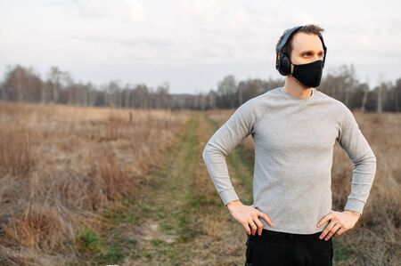 Sports during quarantine, epidemics. A guy in sportswear and a medical mask stands in a meadow, hands in sides. Headphones on a guyの写真素材