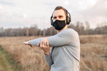 Protect yourself during a pandemic. Sports in a medical mask. Young atletic guy in headphones doing outdoor workout. Closeup portrait in sunset lightの写真素材