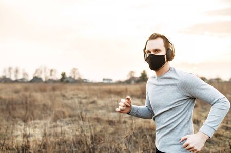 Sports during the quarantine period. A young guy in a black protective medical mask is engaged in jogging, cardio training in the countryside. He is running on a dirt road with headphonesの写真素材
