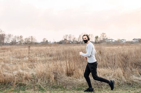 Sport during quarantine, self-isolation in the countryside. A young athletic guy is jogging on a dirt road in the meadow. He is wearing a black medical mask and headphonesの写真素材