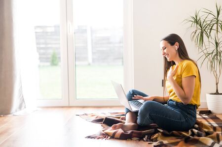 Self-isolation during quarantine. Video communication, zoom. Young positive woman in a yellow casual t-shirt communicates via video communication using a laptop. Side viewの写真素材