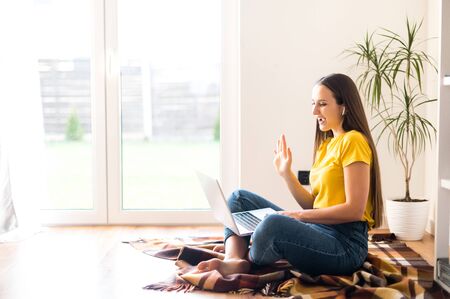 Self-isolation during quarantine. Video communication, zoom. Young positive woman in a yellow casual t-shirt communicates via video communication using a laptop, she waving hello to the screenの写真素材