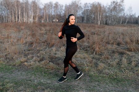 Young attractive athletic woman in a black sports outfit and in headphones jogging, getting ready for a marathon in the countryside. Outdoor cardio training conceptの写真素材
