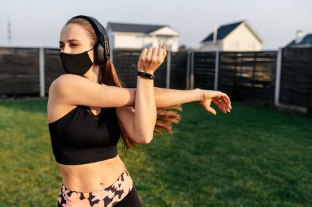 Get in shape during quarantine. Young woman is doing cardio workout in the backyard. She in a medical mask doing stretching on the grass. Spending time with healthy benefit during coronavirus epidemiaの写真素材