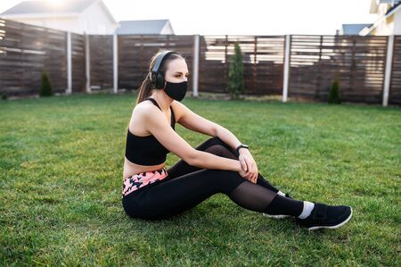 Self-discipline and sports training during quarantine. Portrait of a young attractive woman in a medical mask and in headphones, she is in sportswear in the backyard of the house, outdoors. Total black wearの写真素材