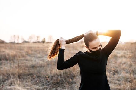 The concept of sports training during quarantine. Young woman in a black sports outfit and in a black medical mask outdoors, she is tying her long hairの写真素材
