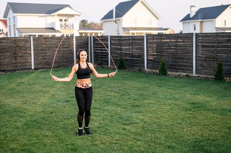Sport lifestyle, healthy lifestyle. Cardio workout in the backyard. A young girl in sportswear is playing with a skipping rope. Weight loss workout at homeの写真素材