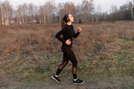 Young attractive athletic woman in a black sports outfit and in headphones jogging, getting ready for a marathon in the countryside. Outdoor cardio training conceptの写真素材