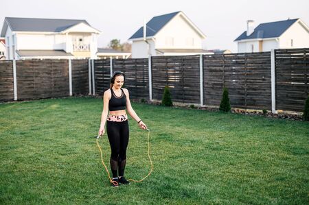Get in shape by summer at home. Young active woman doing cardio workout in the backyard of the house. She is in sportswear and with headphones jumps with a skipping rope the grassの写真素材