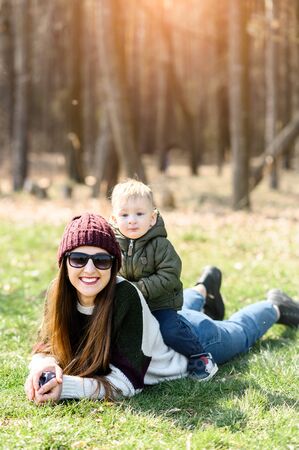 Happy mom and toddler son spend time outdoors. The kid with blond hair sits on the back of his mother, the mother lies on the green grass. Season autumn-spring.の写真素材