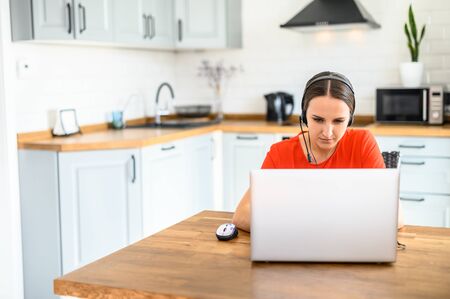 A young business woman uses a headset and laptop computer to work from home while sitting at a table in the kitchen with a serious face. Remote work concept, work from homeの写真素材