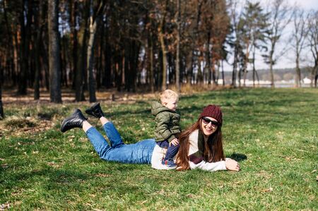 The kid with blond hair sits on his mother's back, mother lying the green grass. Concept of family time together outdoorsの写真素材