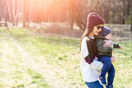 A stylish young mother in a knitted hat and sunglasses carries a baby boy in her arms, they laugh. Family time in the park, in nature, in the forest. Copy paste space for the text on the pictureの写真素材