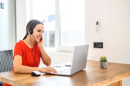 Video conference from home; morning meeting remotely. A young woman uses a headset for online communication, she smiles and talk using a laptop . Work from home conceptの写真素材