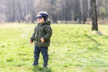 Funny kid stands on the green grass in sunglasses, which are big on him. The boy is in a hat and khaki jacketの写真素材
