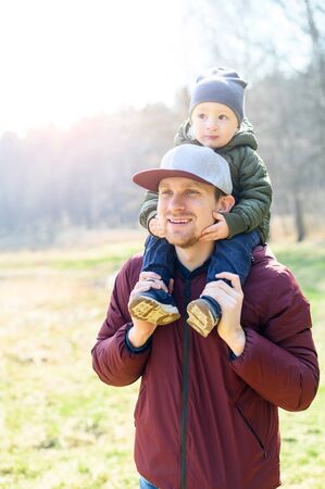 Family time outdoors. A young father and his cute little son in a park together. Dad in cap is carrying his baby boy on shoulders. Vertical closeup photoの写真素材