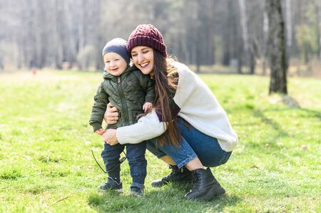 Family time outdoors. Young mother and her cute toddler in spring on the grass in the park. Mom hugs her baby, they are laughing and happy togetherの写真素材