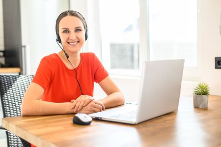 A young business woman uses a headset and laptop computer to work from home while sitting at a table...