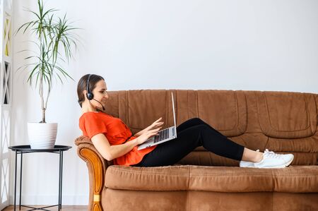 Remote work in a relaxed style. A young woman works from home using hands free headset and laptop, she lies on the sofa, talking and looking at the screenの写真素材