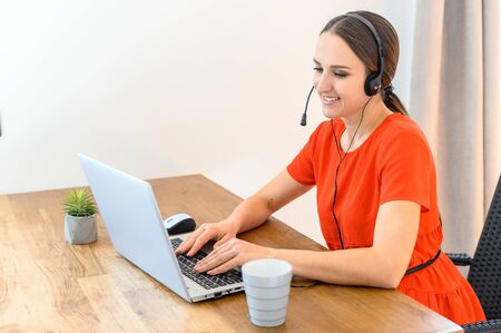 Young female call center operator with hands-free headset talking with client in home office. Concept of remote workの写真素材