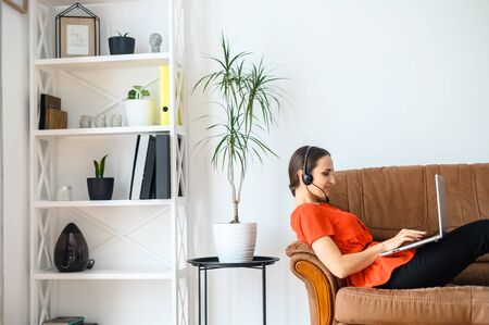 Young woman work from home. She is lying on sofa with a laptop and using headset for online talks. Concept of remote workの写真素材
