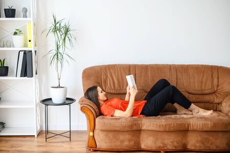 Home leisure, spending time reading books. A young woman is lying on a sofa and reading a book in a cozy home atmosphere.の写真素材